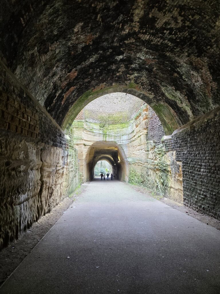A large tunnel carved into rock with natural light coming through a hole in the middle. In the distance, 4 people can be seen walking through the tunnel