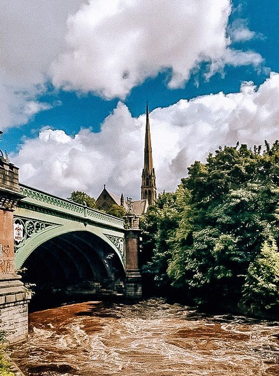 fast flowing river under a bridge with church spire in the background