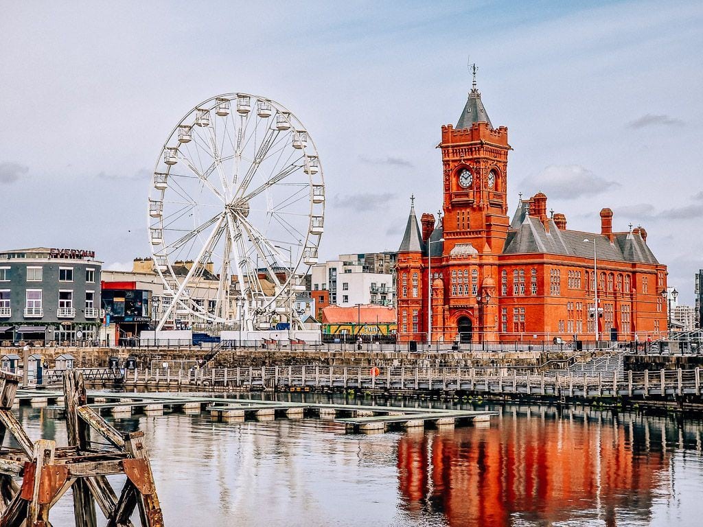 The shore of Cardiff Bay lined with many buildings including a large white Ferris wheel and a massive orange brick building with a clocktower