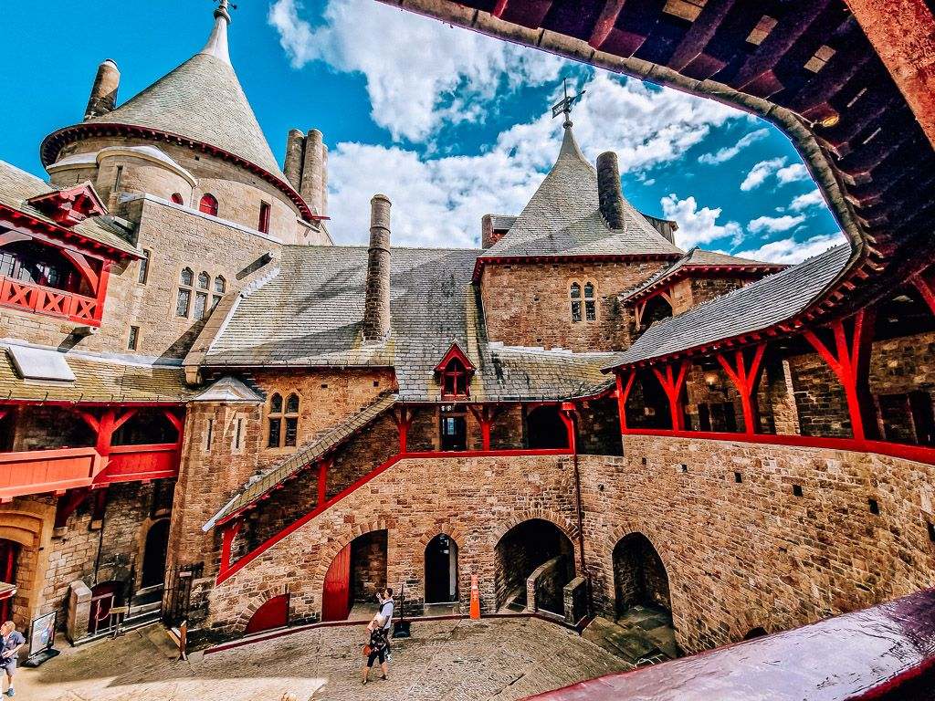 Looking down on a castle courtyard from a balcony, castle is made of dark stone with red wood structure. There are two turrets seen and people walking about below