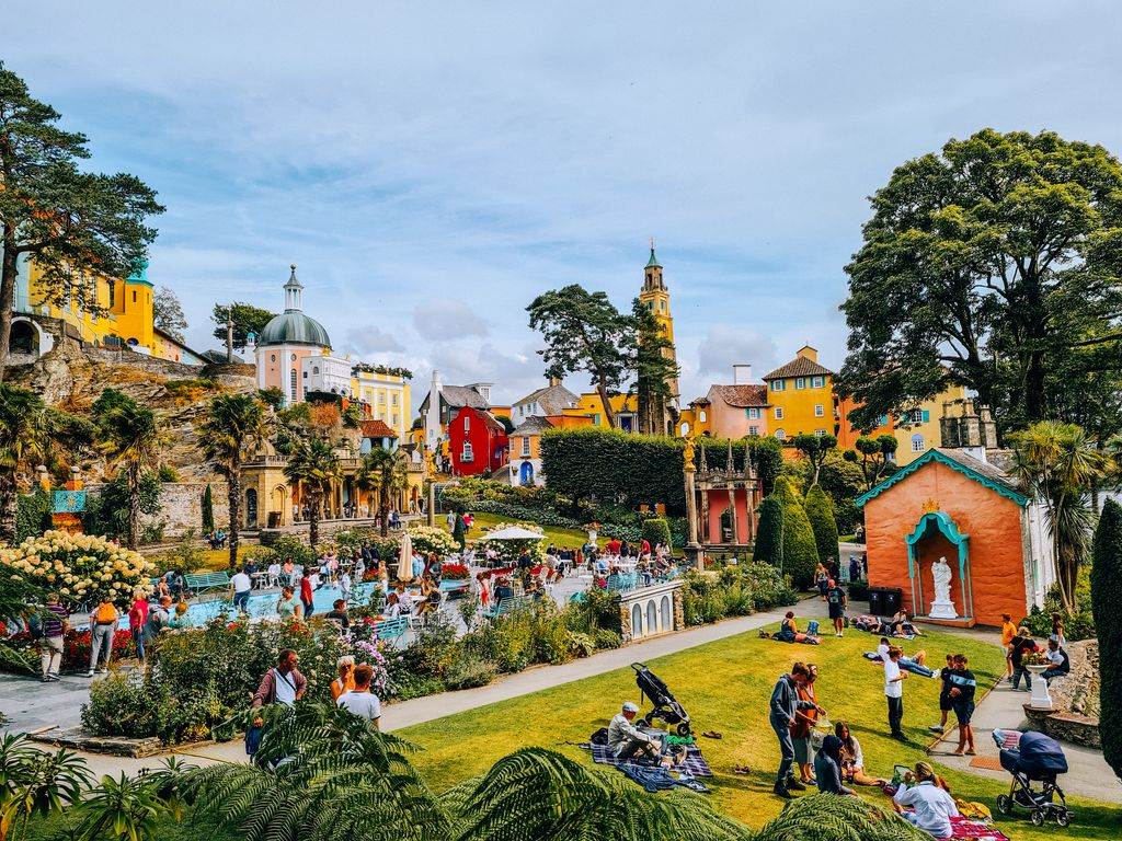 A vibrant image of the italianate village of Portmeirion, with orange, red and yellow buildings and lots of lawn and greenery with people picnicking around a lido where children are playing