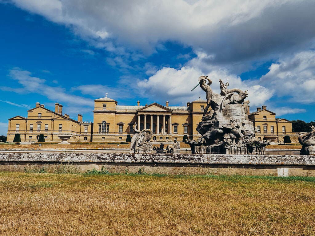A large fountain with many stone statues at its centre and very large yellow stone stately home is in the background