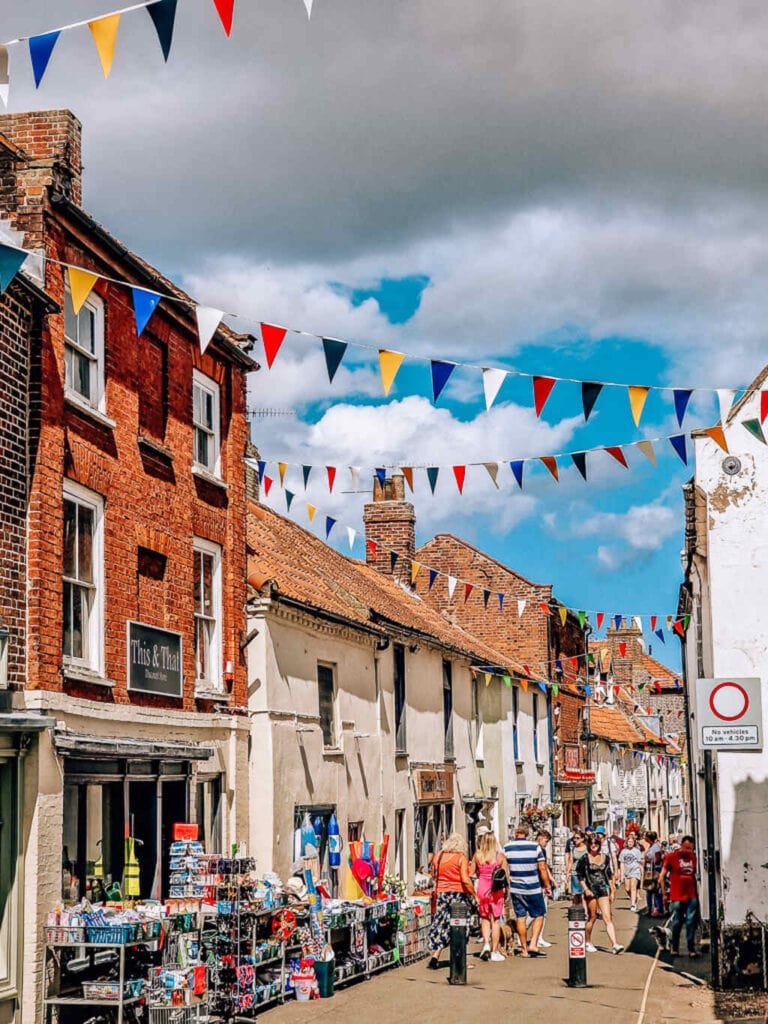 A busy pedestrianised street with many old white and brick shops. Multi-coloured bunting is stringed across the street