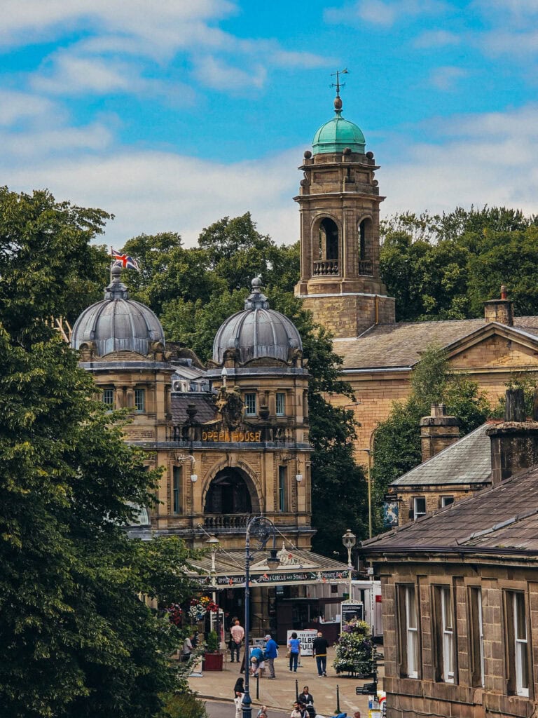 Layers of grand, sandstone buildings. The one at the front is a theatre with two metal domes and an archway above the foyer. Behind is a tower with a turquoise blue dome. Trees surround the picture