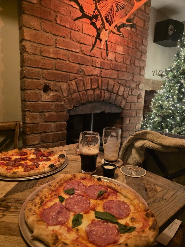 Two pizzas and two half-finished pints of Guinness on a wooden table in a pub. Behind the table is a brick fireplace