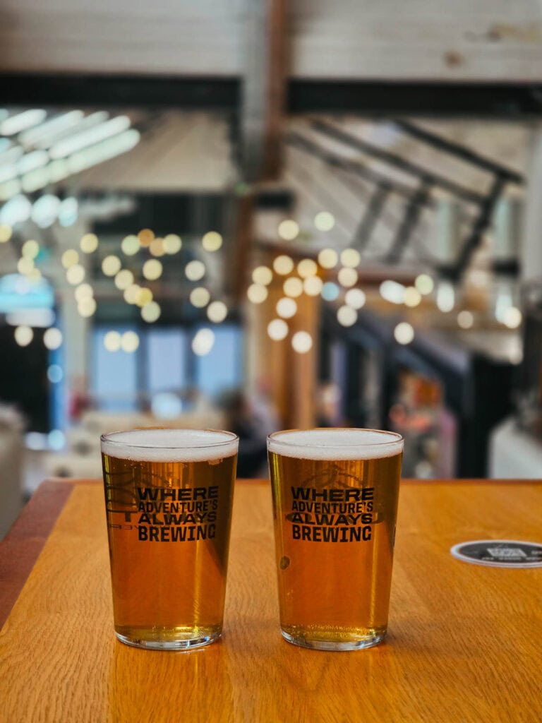 Two full pints of beer on a wooden table, the background is out of focus with fairy lights on the ceiling of an industrial style room.