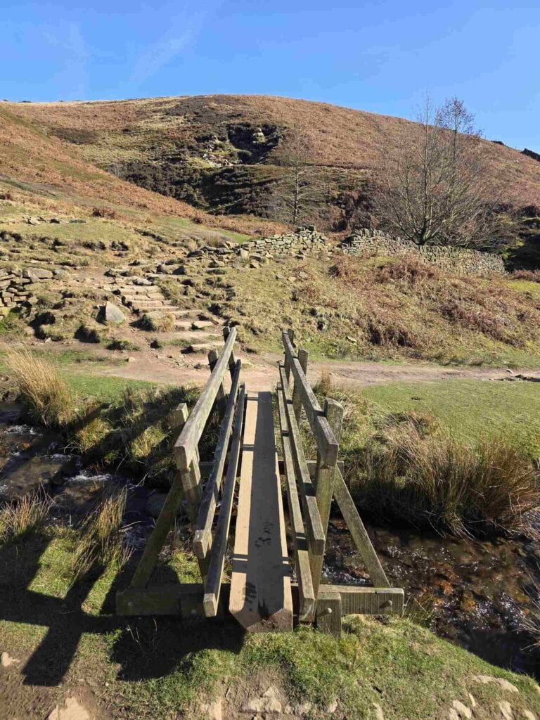 A small wooden footbridge crossing over a small brook leading to stone steps ascending up the hillside beyond. It's a clear day with blue sky.