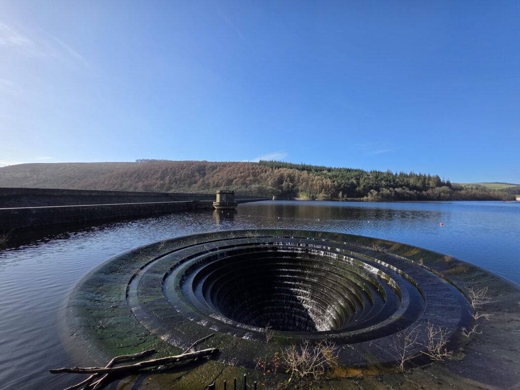 A calm reservoir on a sunny day with a large spillway close to the camera that looks like a plughole with water overflowing from the reservoir into it.